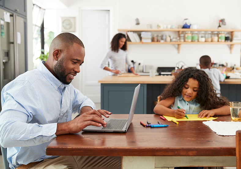Young black man working on laptop in the kitchen with family nearby working on different projects